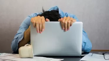 Personne appuyée sur un ordinateur portable, entourée de papiers et lunettes.