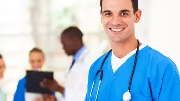 Professionnel de santé souriant en tenue médicale bleue, avec collègues en arrière-plan.