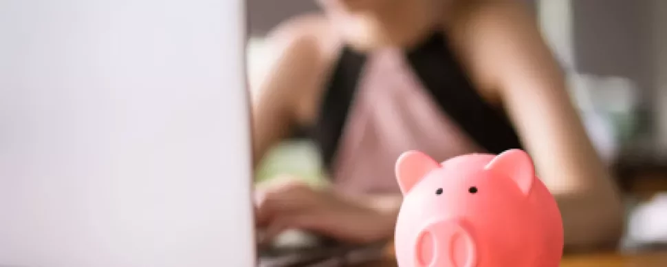 Person typing on laptop with piggy bank in foreground.